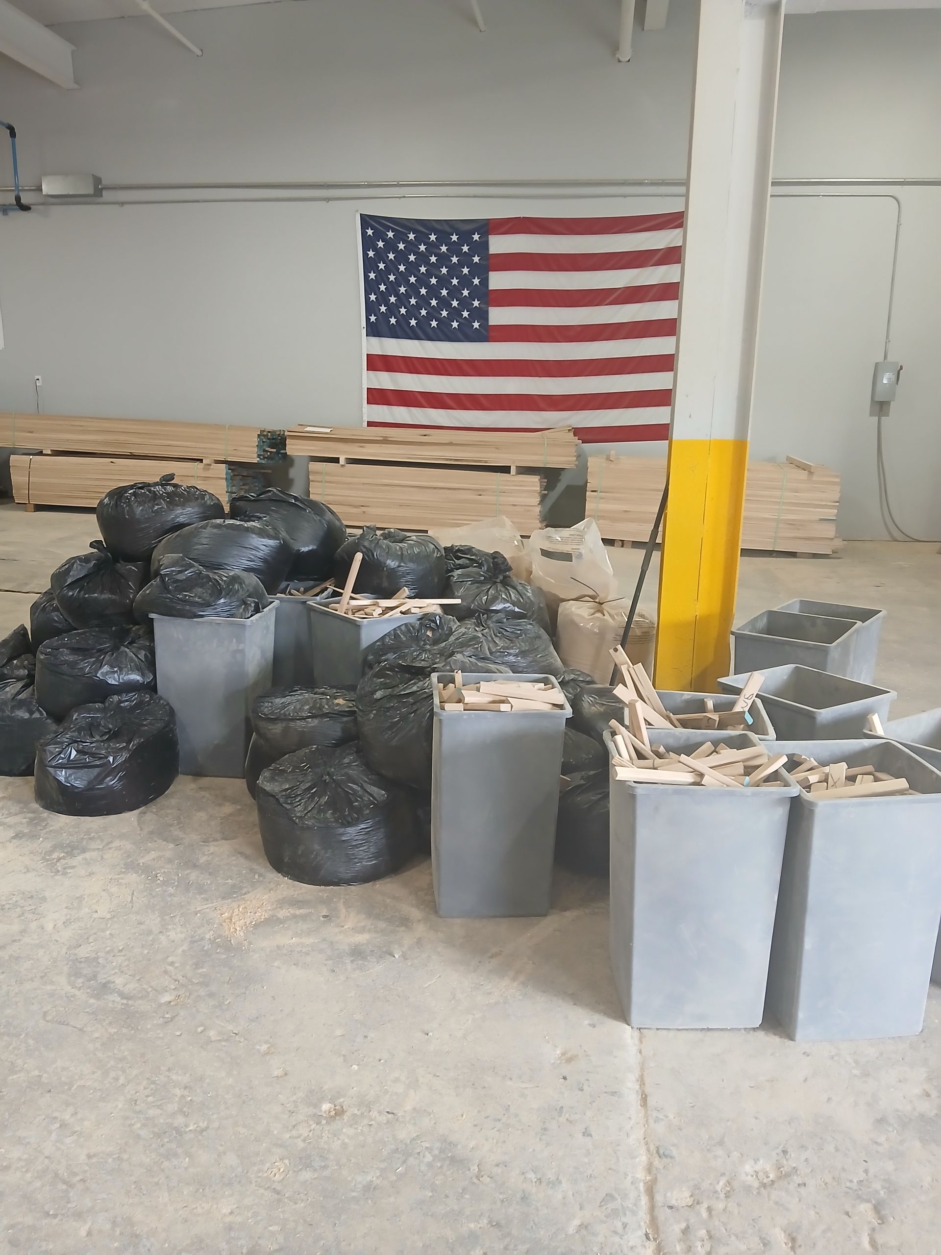 Stacks of black trash bags and gray bins in a warehouse, with an American flag on the wall.