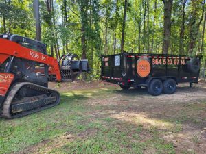 Orange excavator beside a black utility trailer in a wooded clearing.