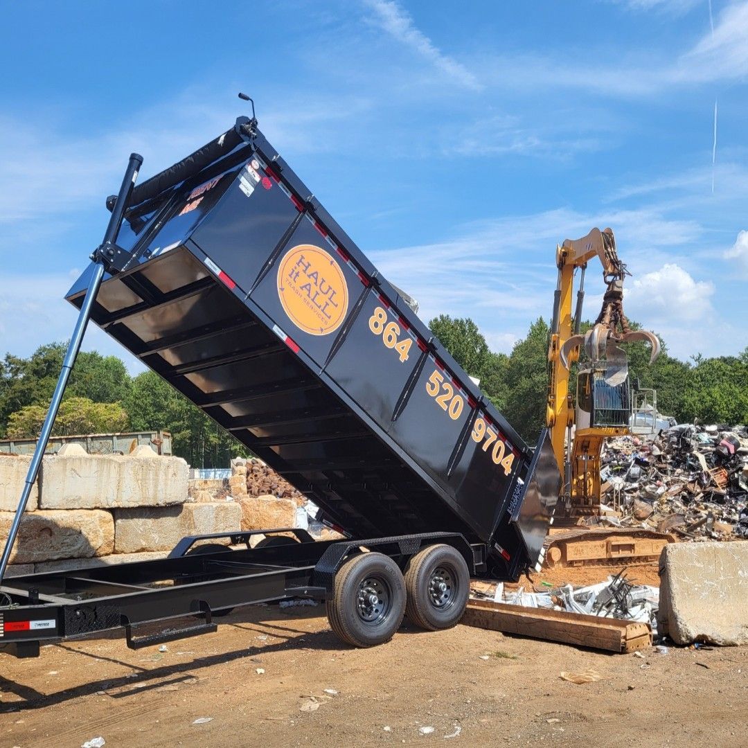 Black dump trailer raised at a scrapyard, unloading debris near an excavator under a blue sky