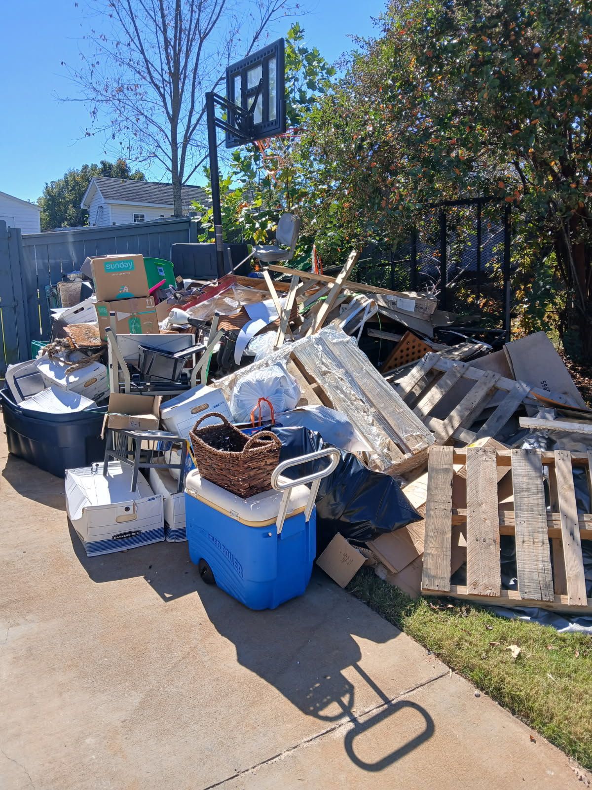 Overflowing trash and debris piled along a sidewalk beside a fence on a sunny day.
