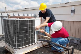 Two workers in hard hats repair an outdoor air conditioning unit on a rooftop.
