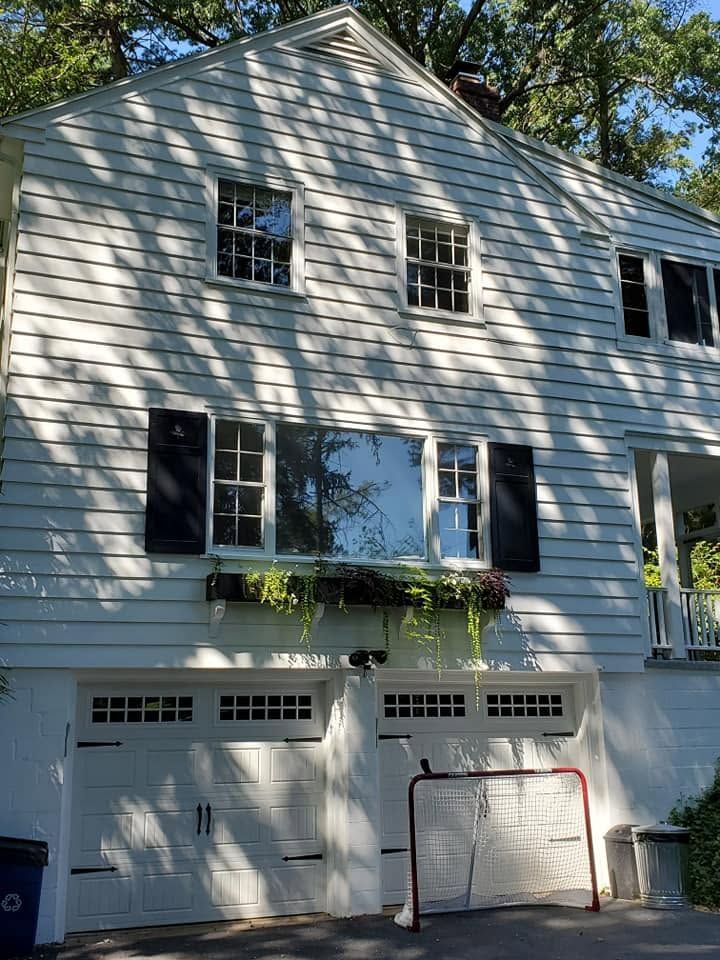 White house with black shutters and garage doors, seen from the front with tree shadows across the siding.
