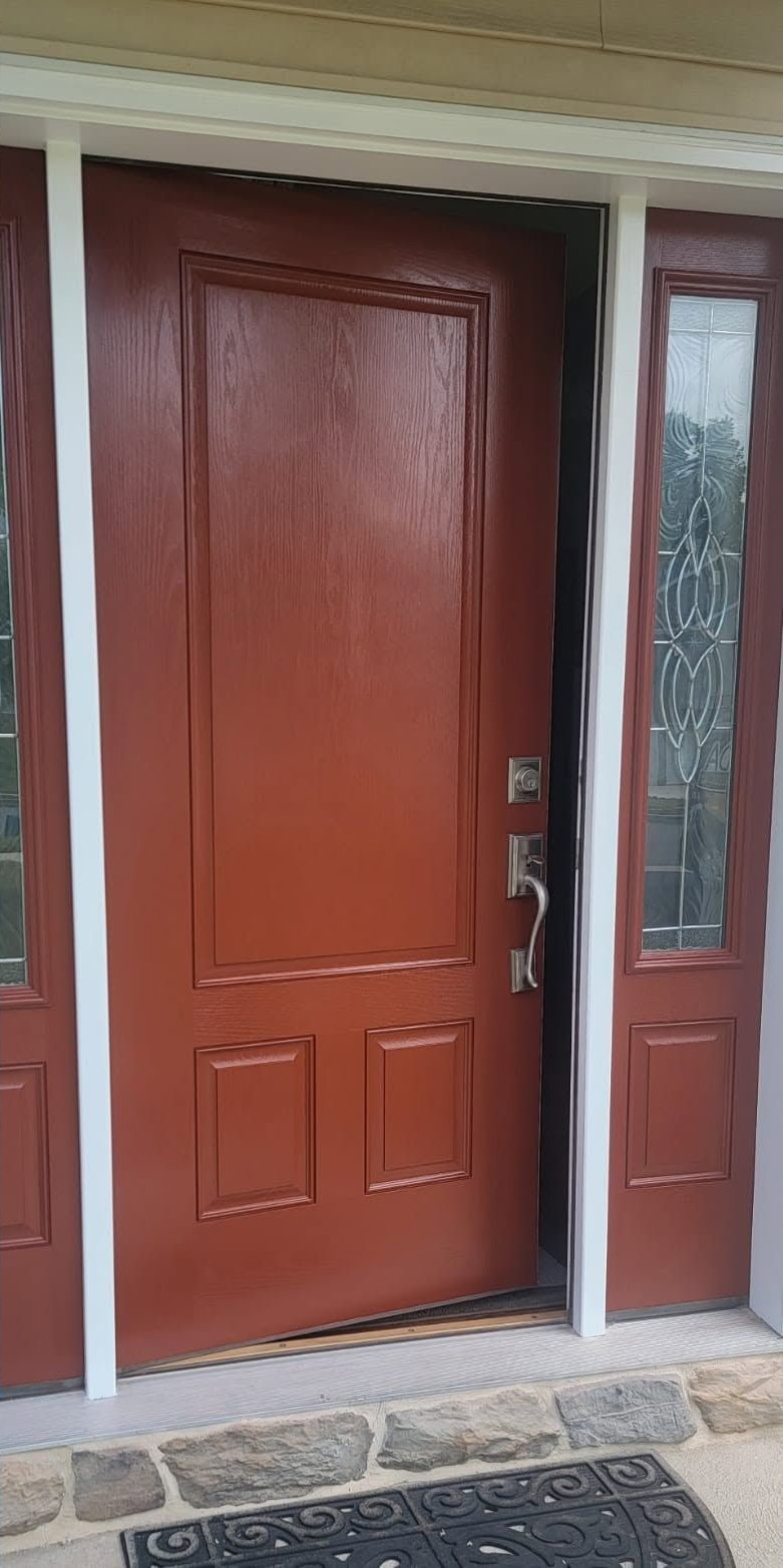 Red front door with decorative panels beside a narrow sidelight window and stone threshold.