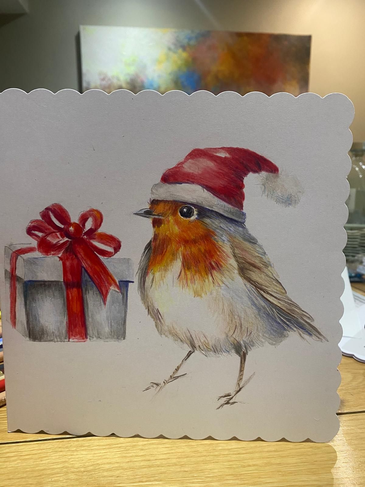 Christmas card: robin wearing a Santa hat, next to a gift box with red ribbon, white background.