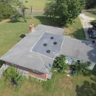 Aerial view of a gray-roofed house with a central flat section containing tires, surrounded by green grass and trees.