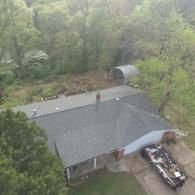 Overhead view of a house with a gray roof and a driveway. A wooded area surrounds the home.