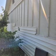 Exterior of a white house with peeling paint and vertical siding. Sunlight illuminates the building.