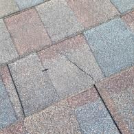 Close-up view of asphalt shingles on a roof; gray, red, and blue tones, with one shingle cracked.