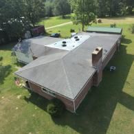 Aerial view of a long brick building with a dark gray roof, chimney, and surrounded by green grass and trees.