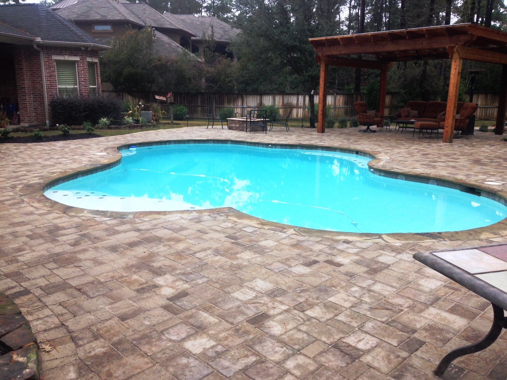A large swimming pool is surrounded by a brick patio and a pergola.