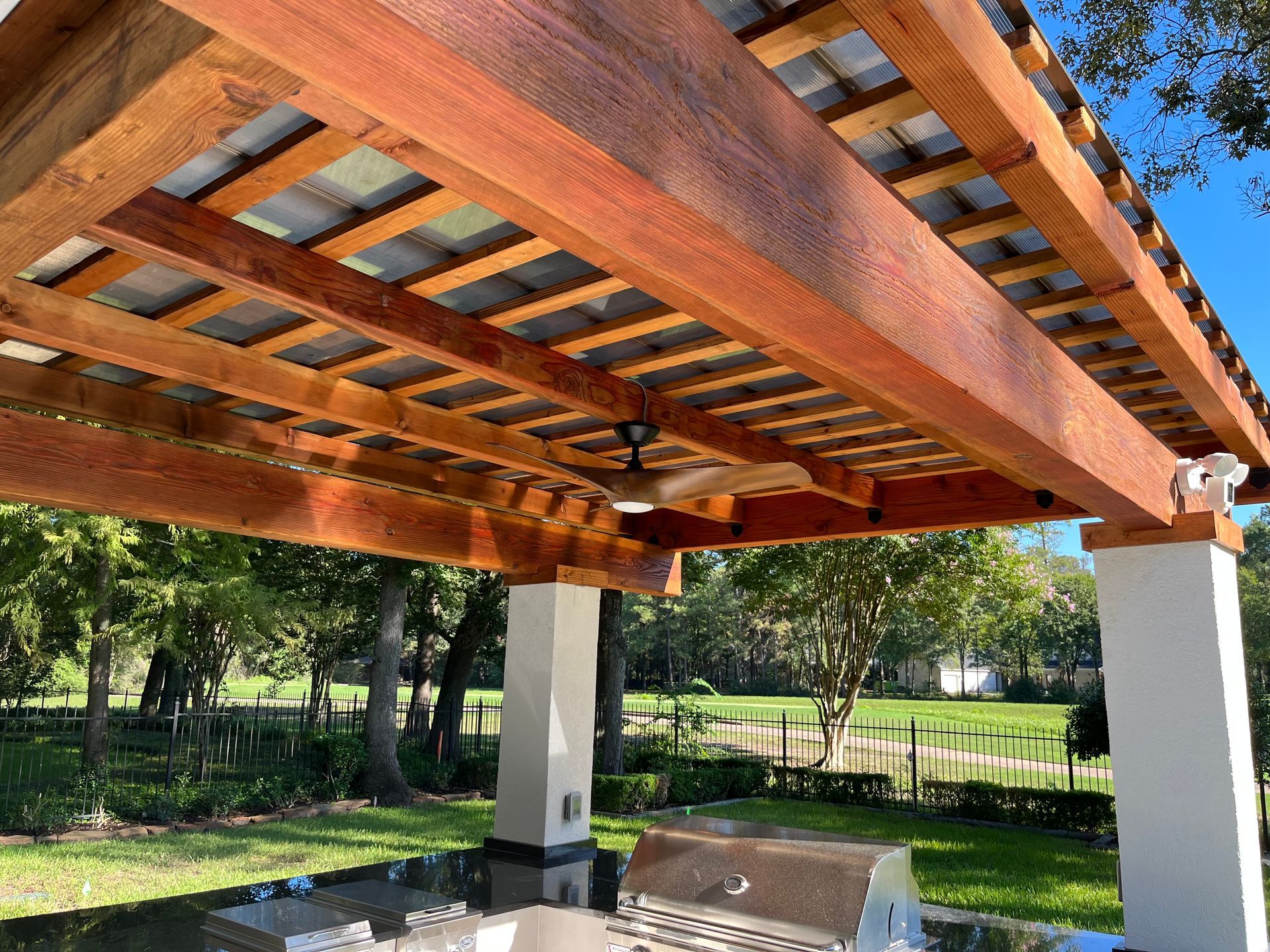 A wooden pergola with a ceiling fan and a grill underneath it.