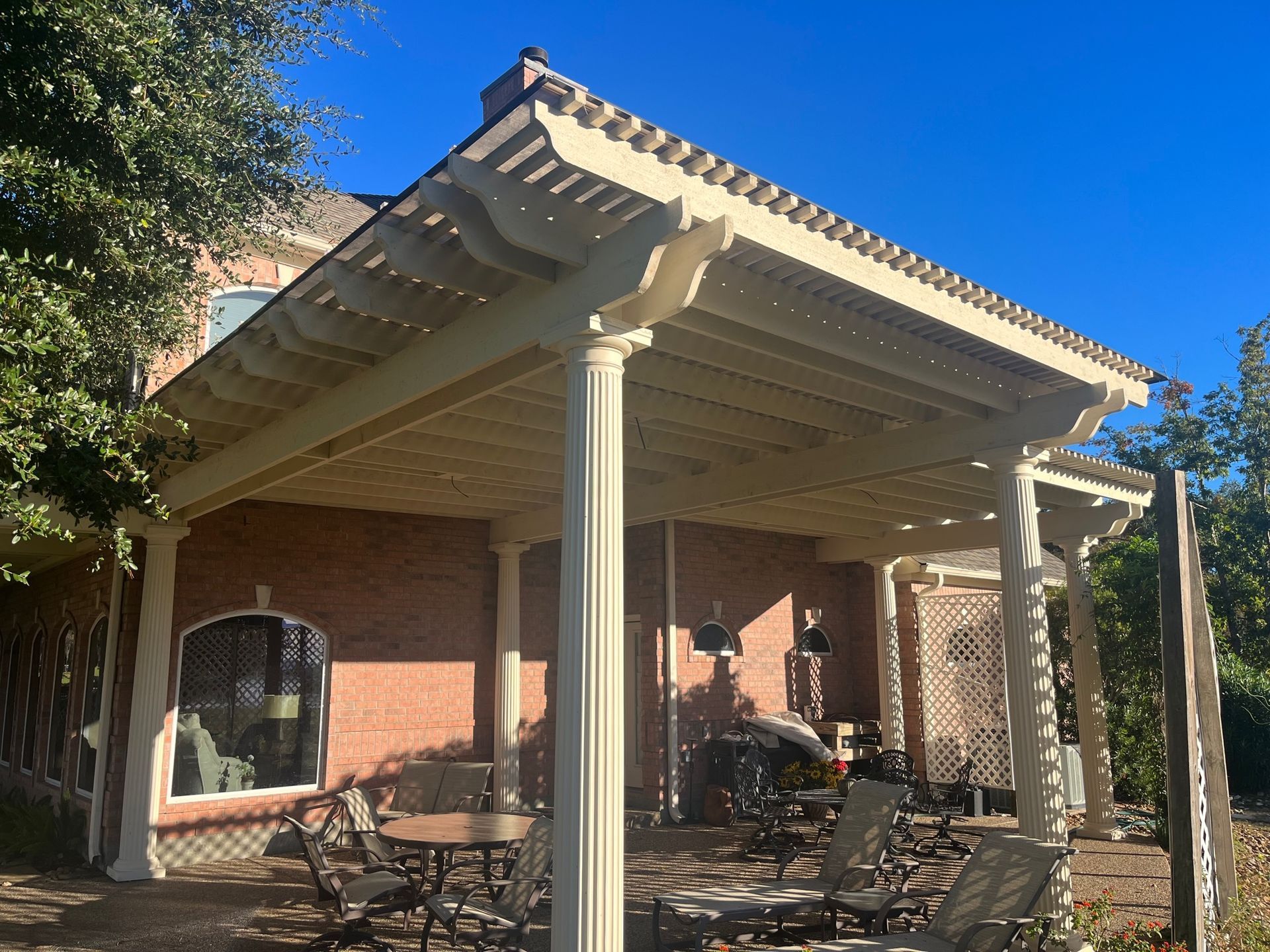 A white pergola with tables and chairs underneath it in front of a brick house.