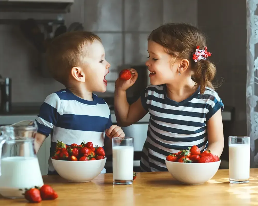 Un niño y una niña están sentados en una mesa comiendo fresas y bebiendo leche.
