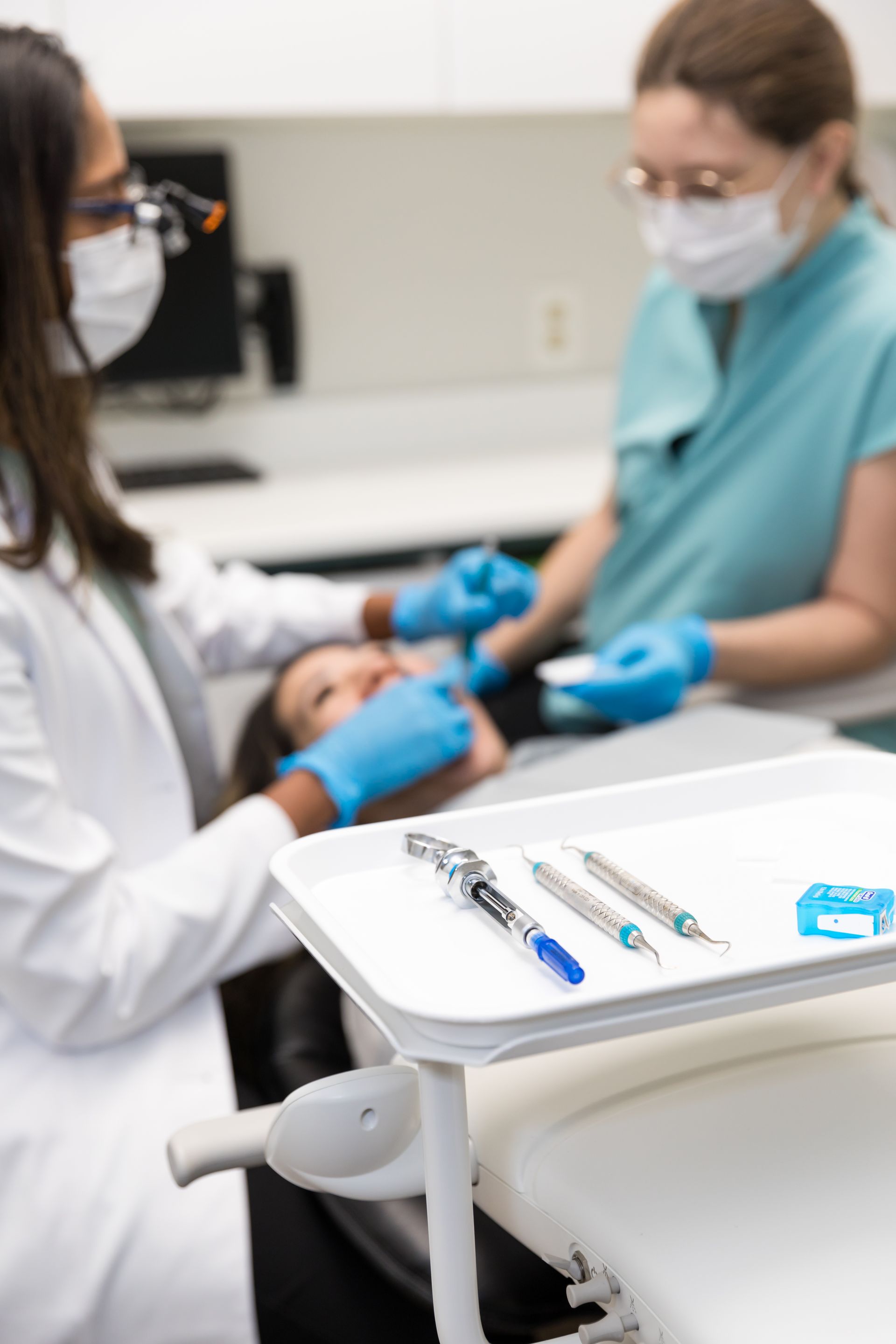 Dentist and assistant treating a patient in an examination room; dental tools visible on a tray.