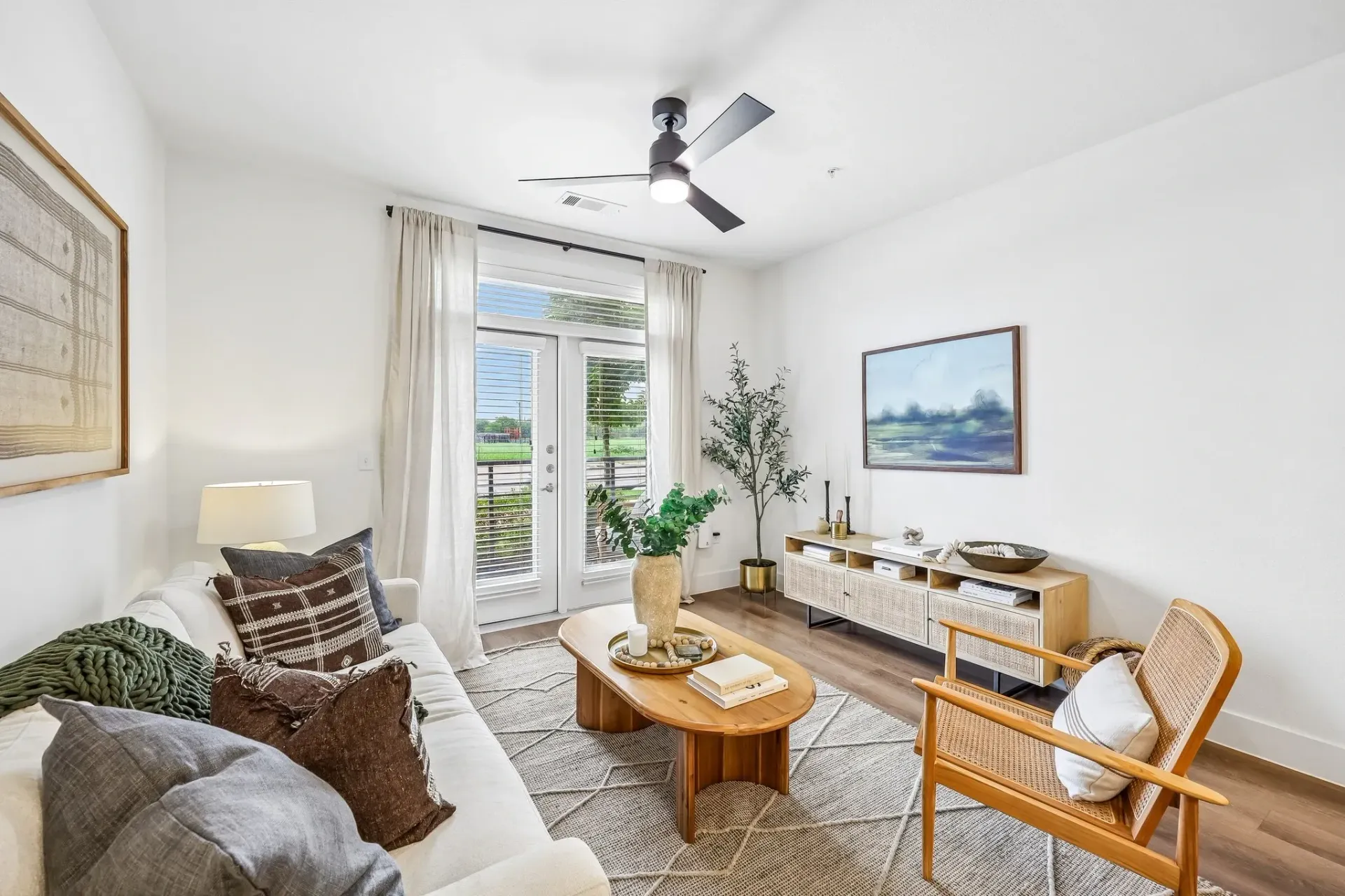 Modern living room with a white sofa, coffee table, and TV stand, featuring large windows and a ceiling fan.