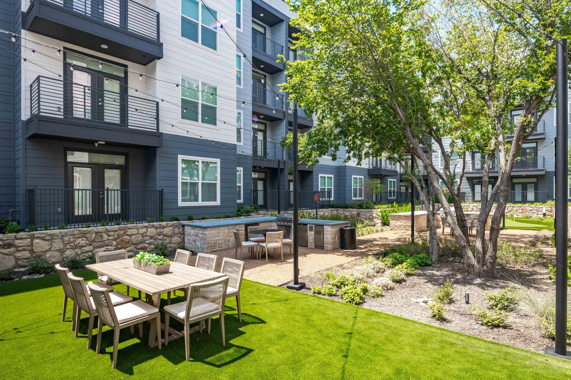 Outdoor dining area with a large table and chairs on artificial turf, adjacent to an apartment building.