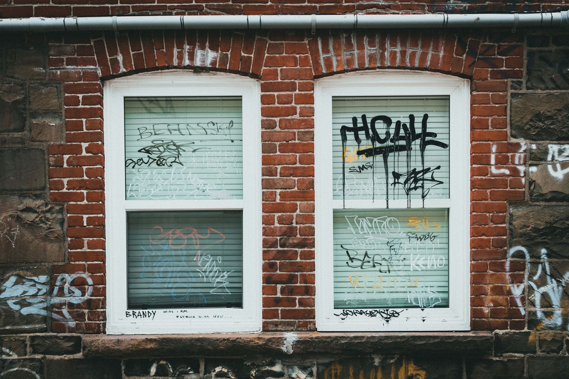 Brick wall with two white-framed windows covered in graffiti and weathered blinds