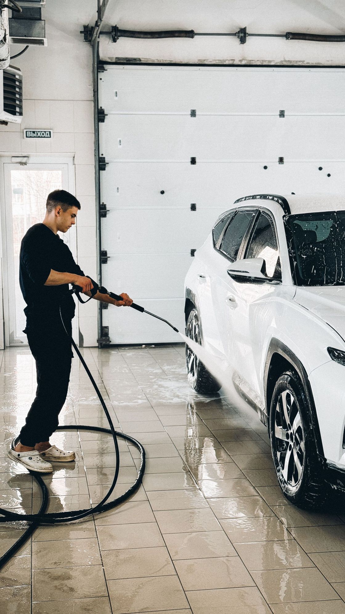 Person pressure-washes a white SUV in a garage with soapy water on the floor