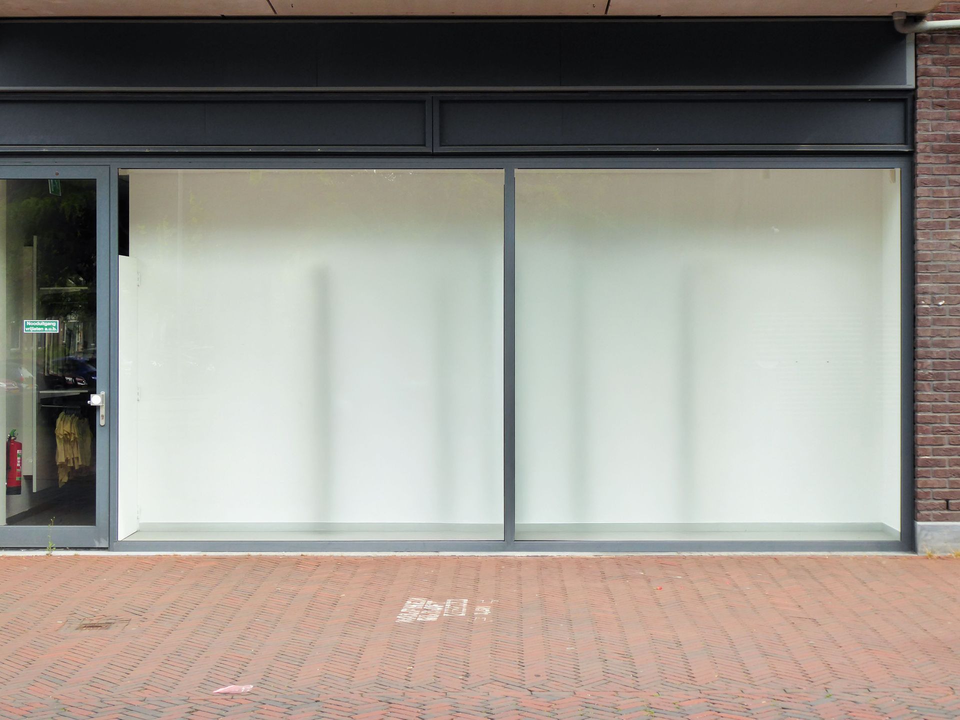 Empty storefront with large white display windows and a black awning above a brick sidewalk.