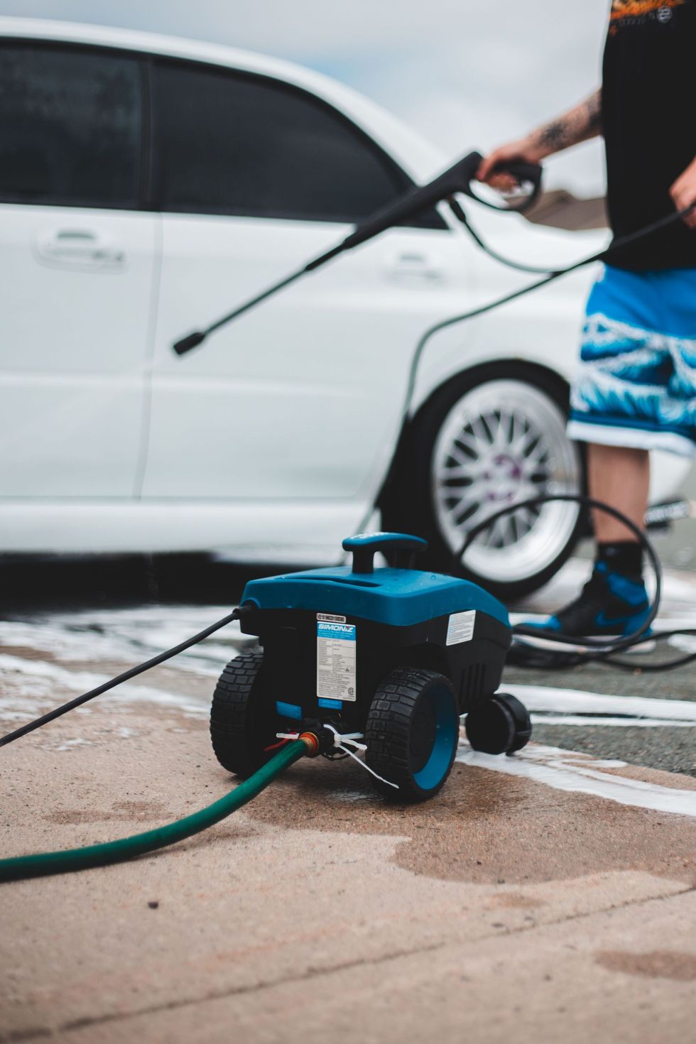 Person pressure-washing a white car with a blue-and-black hose reel on a wet driveway