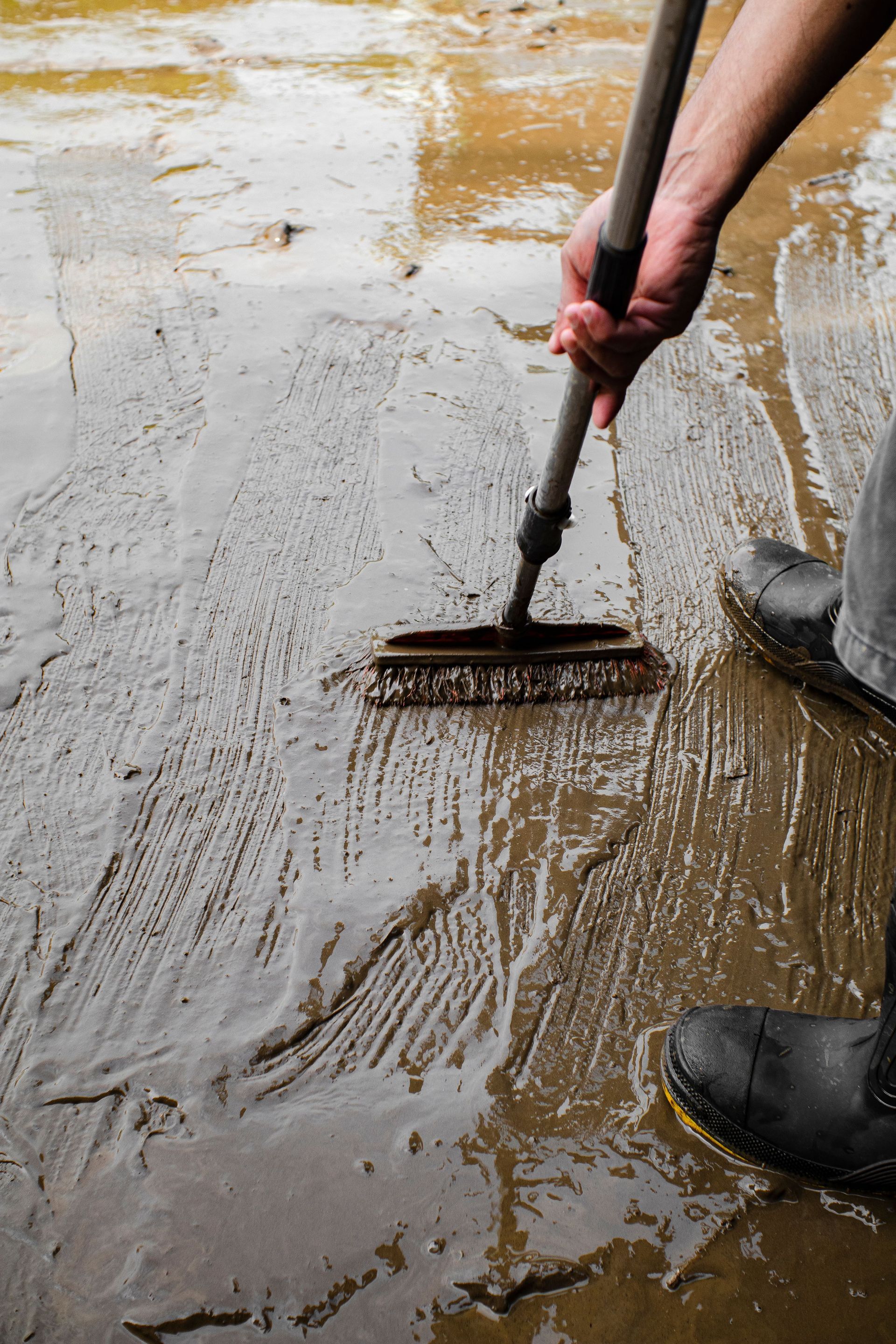 Person raking muddy water off a wet floor with a long-handled broom, boots visible