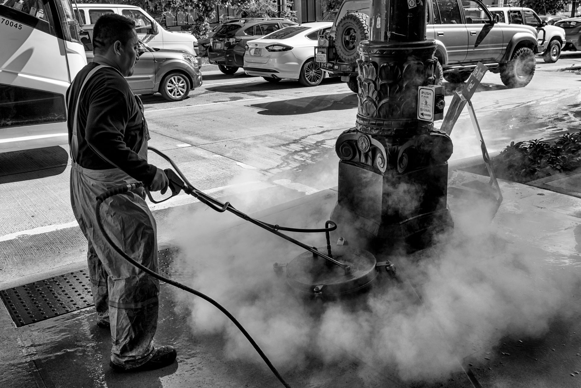 Worker pressure-washes a steam-covered sidewalk beside parked vehicles in black and white.