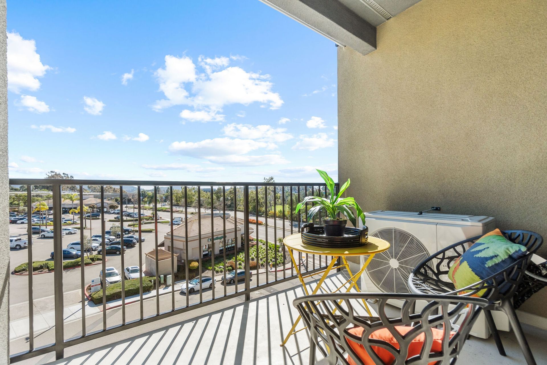 Balcony with small table, black chair, air conditioner, and potted plant, overlooking a parking lot on a sunny day.