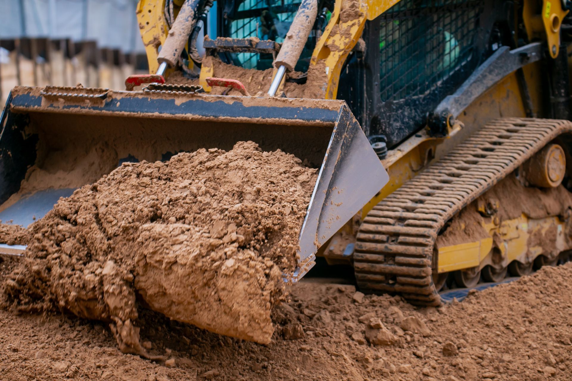 Orange excavator digging beside a road under a blue sky, dirt mound in foreground, trees in background.