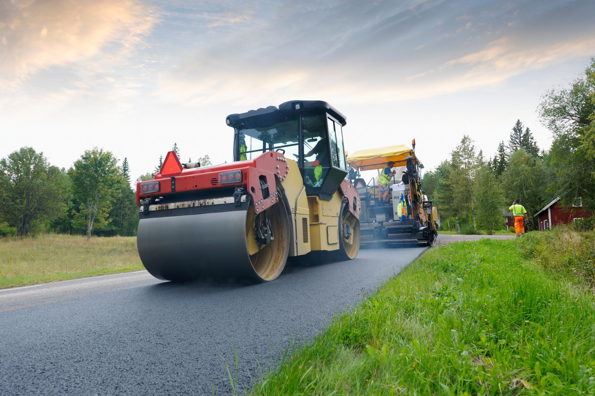 Road paving in progress, a steamroller compacting fresh asphalt on a road, with a paving machine and workers in a rural setting.