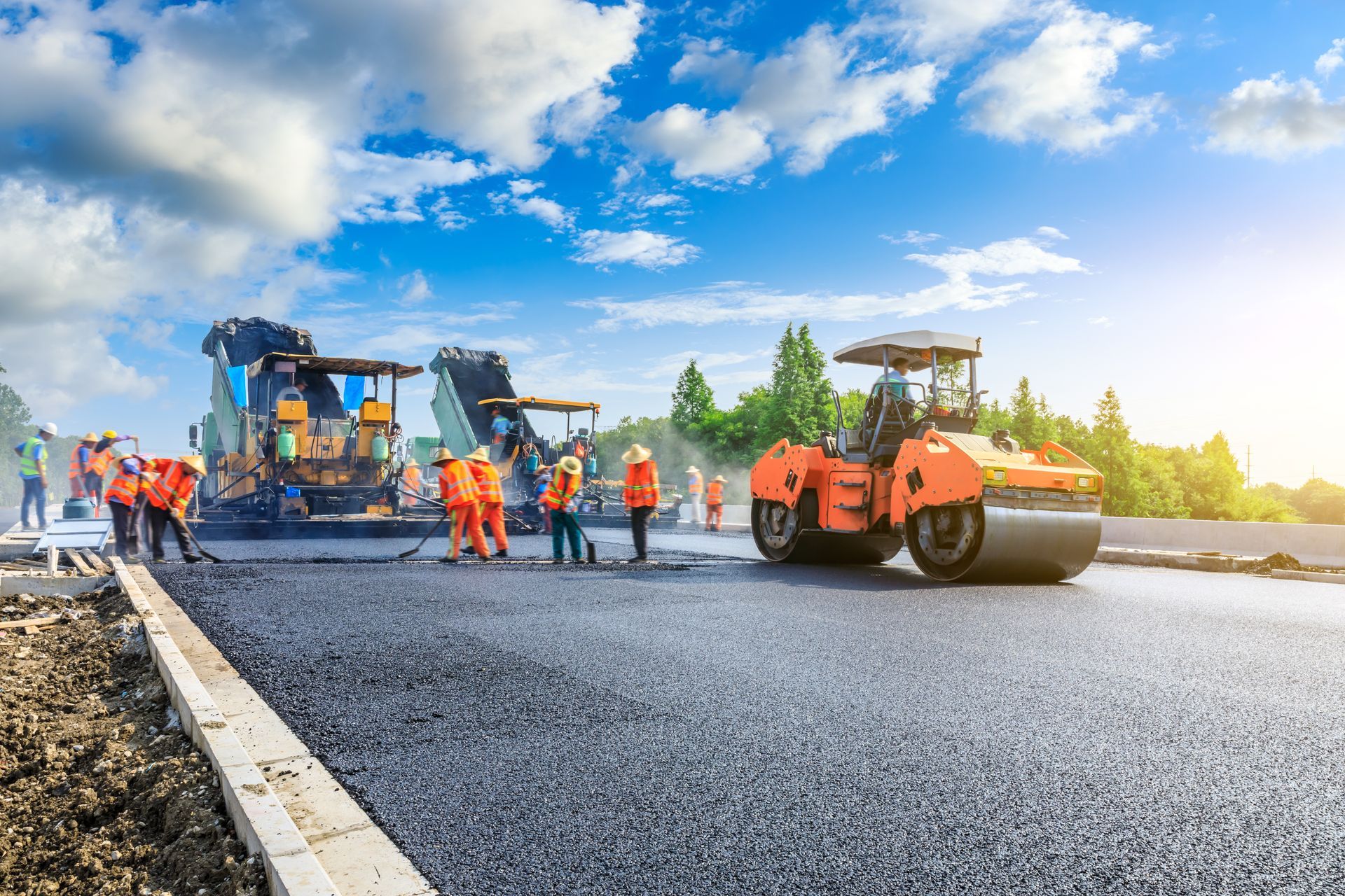 Road construction: Asphalt being laid by machinery, workers in orange vests. Sunny day, blue sky.