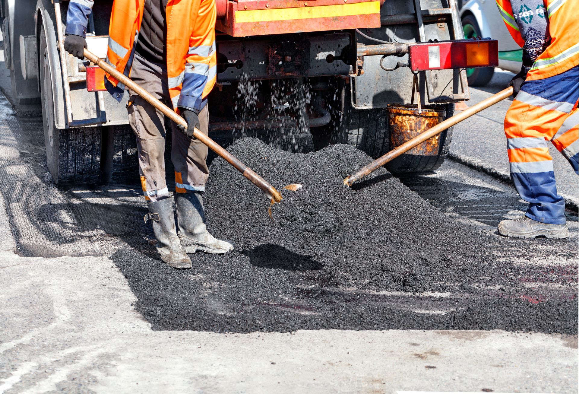 Road workers in orange vests spreading asphalt with rakes near a truck.