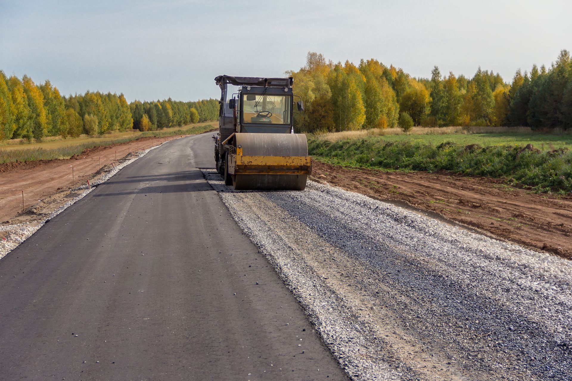 Road roller compacting asphalt on a newly paved road. Gravel and trees are visible in the background. Road roller compacting asphalt on a newly paved road. Gravel and trees are visible in the background.