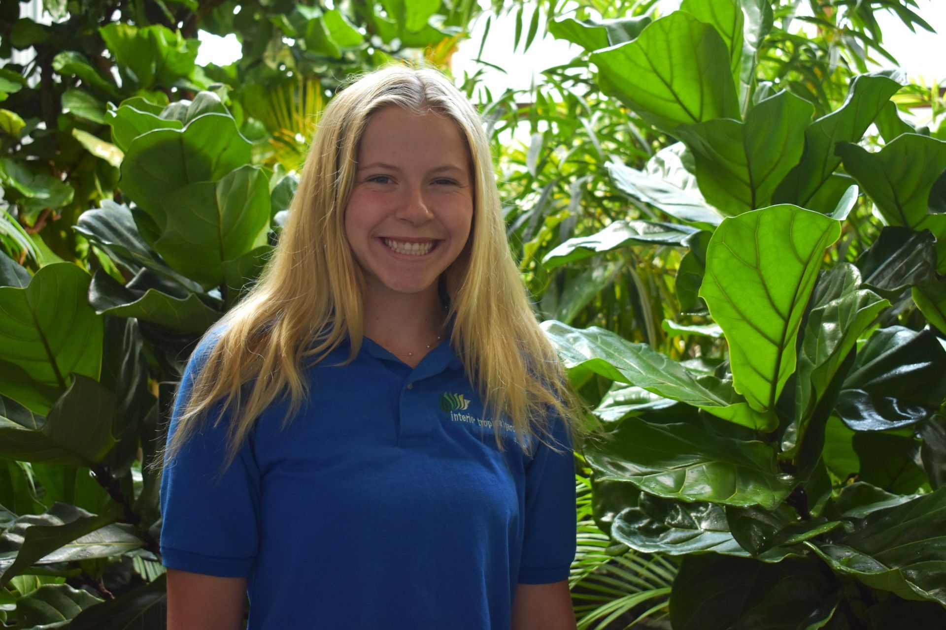 Blond woman smiling in a blue shirt stands in front of large green leaves.