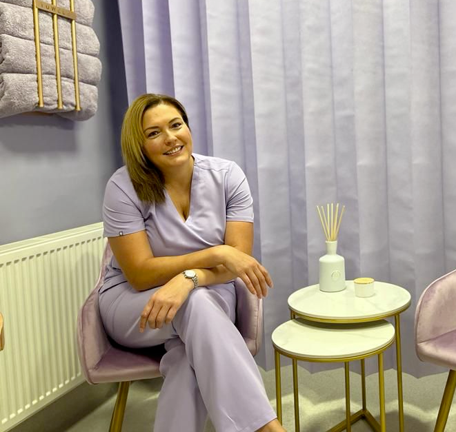 Woman in lavender scrubs smiles, sitting in a pink chair. Room features lavender decor and small side table.