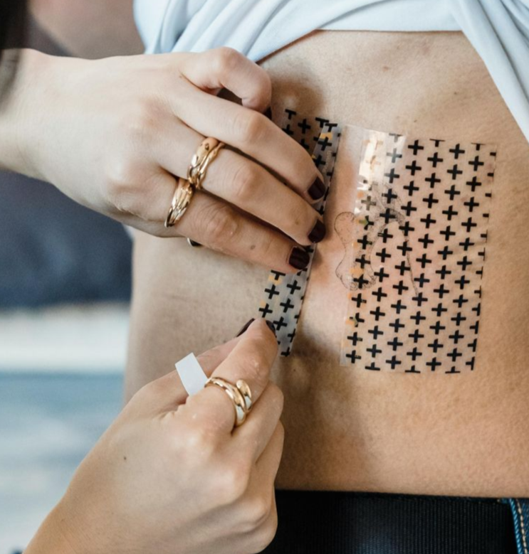 Person applying a bandage with black plus signs to a torso. Hands wear rings; the person is indoors.