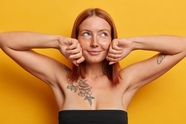 Woman in black top with arms raised, hands near face, looking upwards. Yellow background. Tattoos visible.