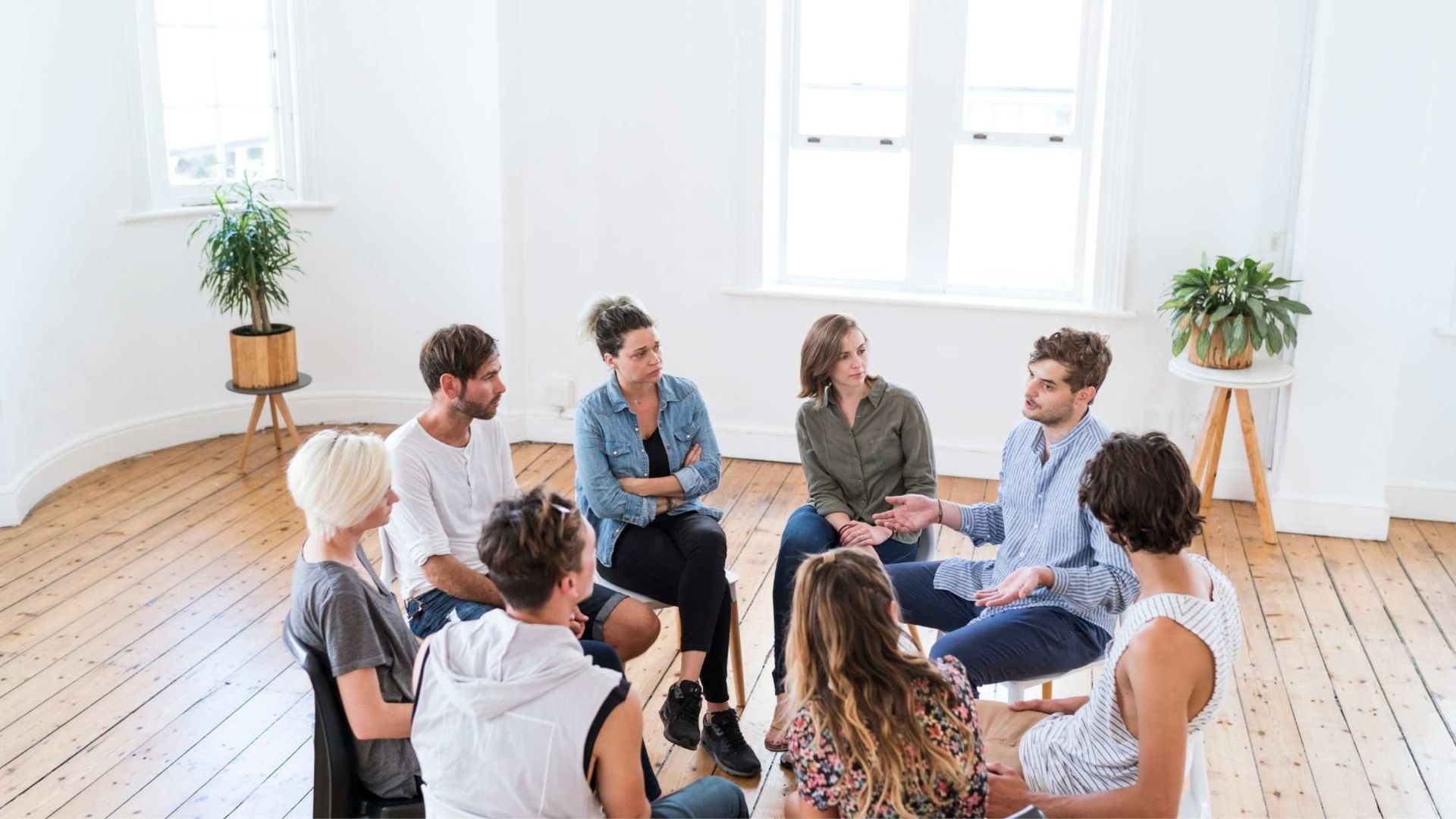 A group of people sit in a circle on a wooden floor in a bright, minimalist room, engaged in a discussion.