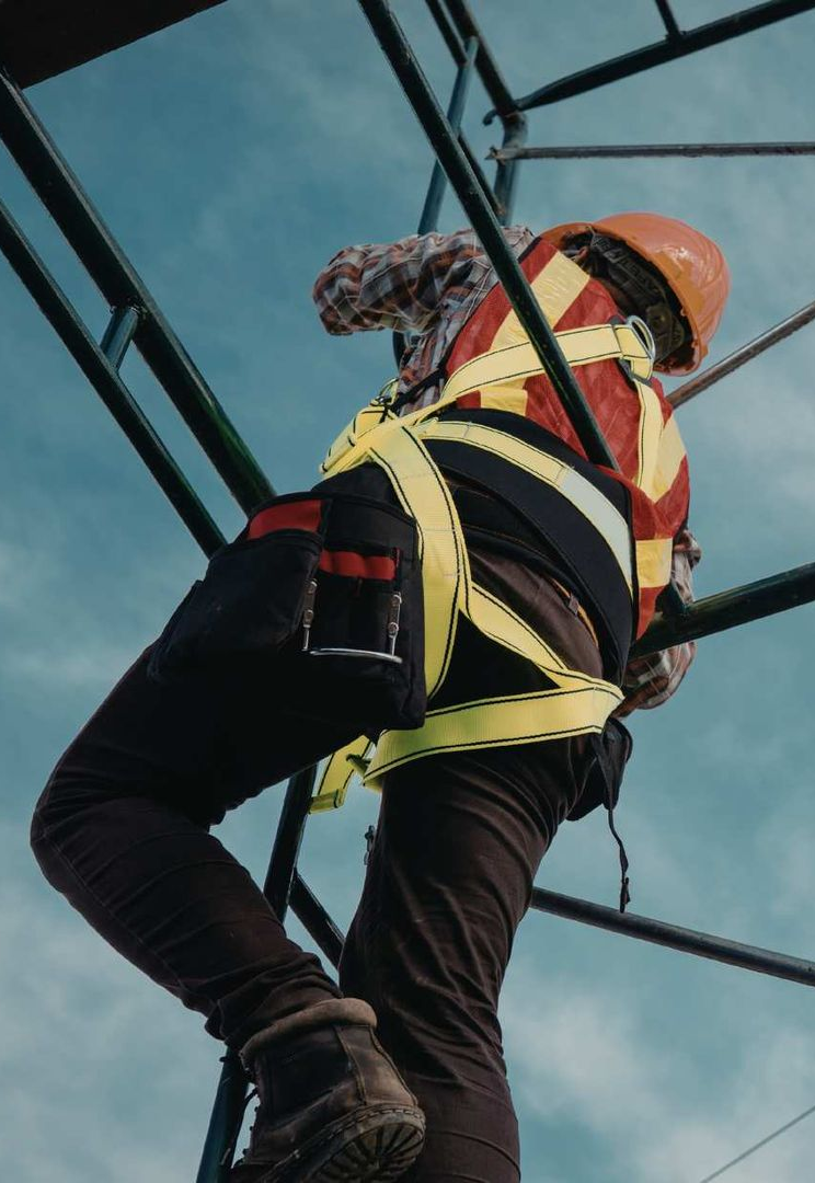 A worker in a high-visibility harness and hard hat climbs a steel construction frame against a blue sky.