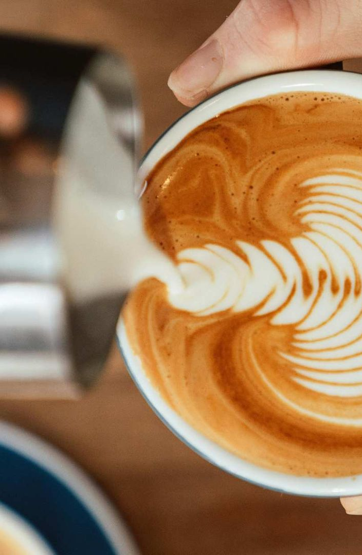 A close-up of a hand pouring steamed milk into espresso to create a white leaf-shaped latte art design.