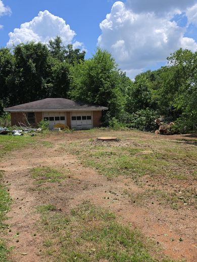 Small brick house behind a cleared dirt lot, framed by tall green trees under a bright blue sky.