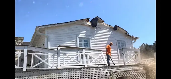 Person in an orange shirt on a porch beside a damaged white house with roof debris.