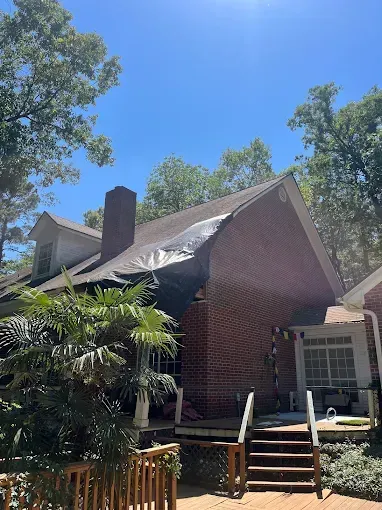 Brick house with damaged roof and chimney, viewed from a wooden stairway among trees.