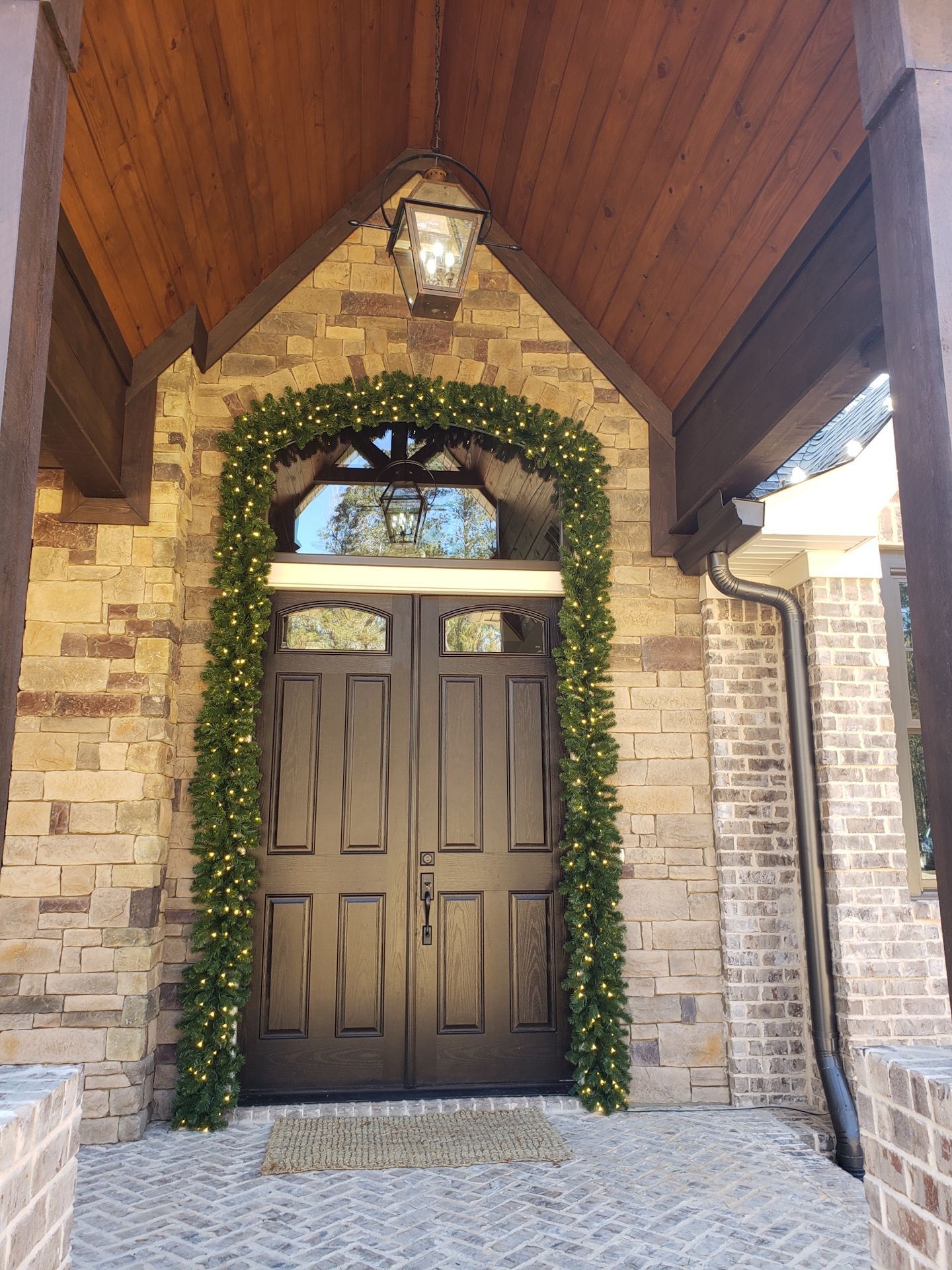 Stone entryway with dark double doors, arched ivy garland, and a hanging lantern under a wooden porch ceiling
