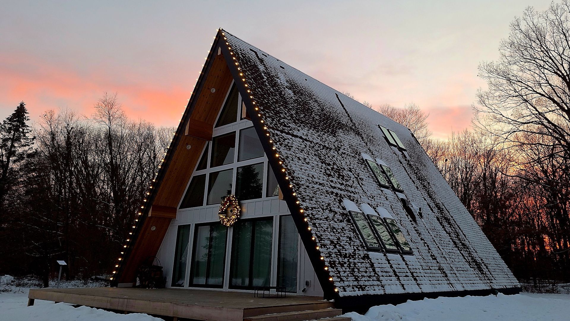 A-frame cabin with snow on the roof at sunset, surrounded by bare winter trees.