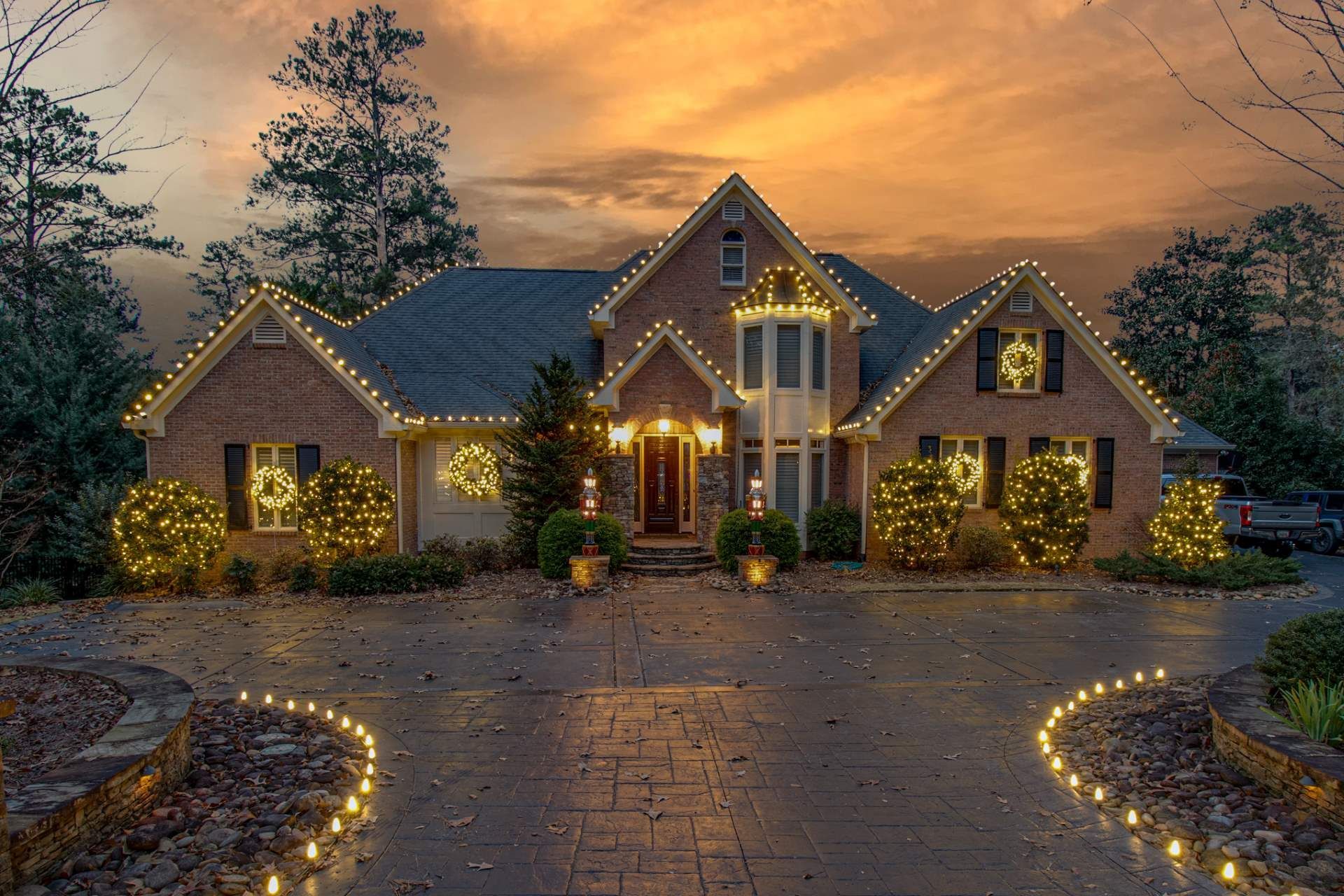 Brick house at dusk with warm holiday lights, stone driveway, and tall trees under an orange sunset sky