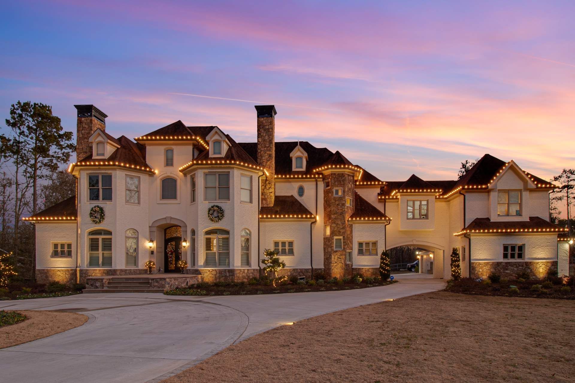 Large cream-colored luxury mansion at dusk with warm lights, arched windows, and a curved driveway under a pink sky