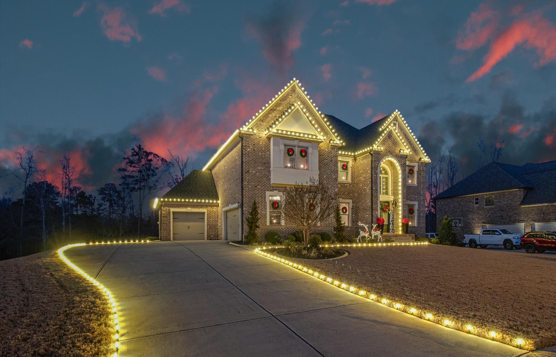 House outlined with warm white lights at dusk, with a lit driveway and cloudy sky.