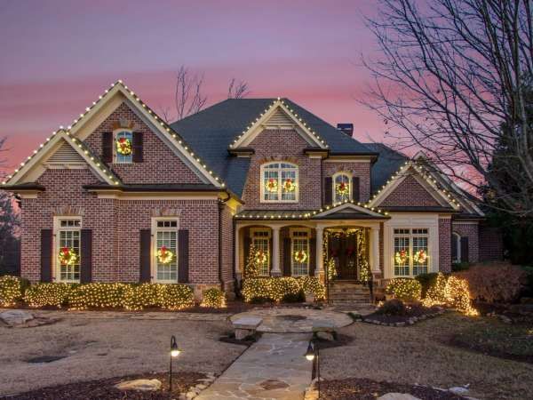 Large brick suburban house with lit windows and holiday lights at dusk, under a pink sky