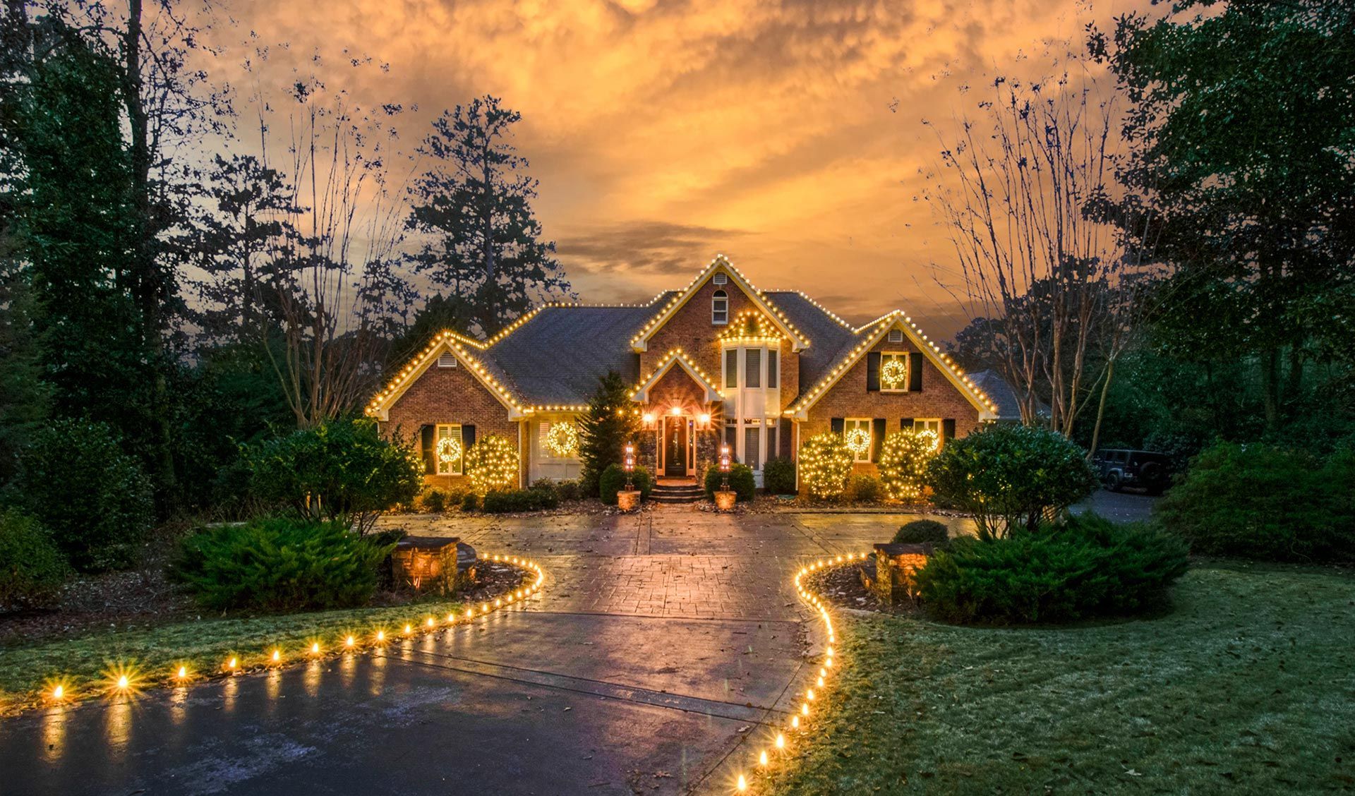 Luxurious house with warm lights and illuminated driveway at dusk under a dramatic orange sky