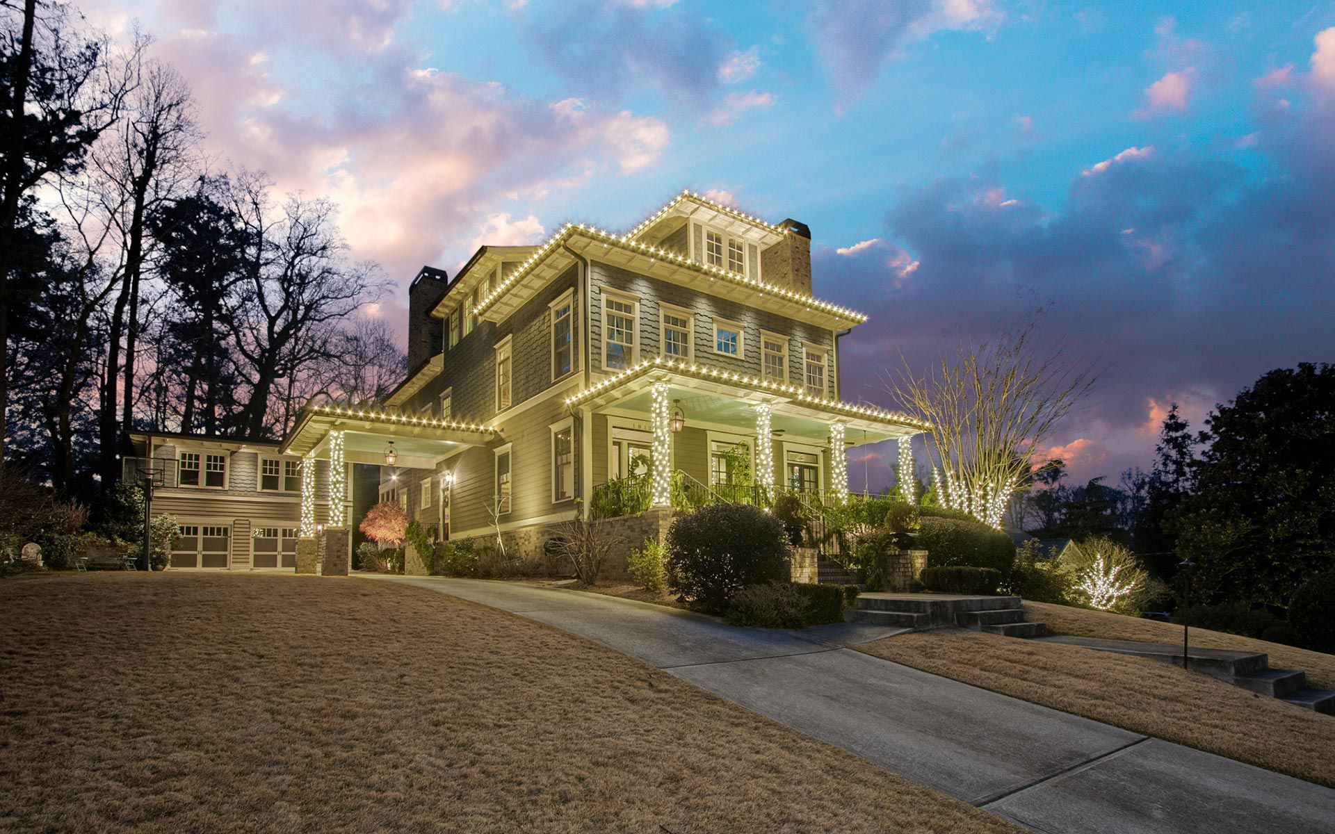 Large illuminated house on a hill at dusk, with lit windows, trees, and a driveway leading up to it.